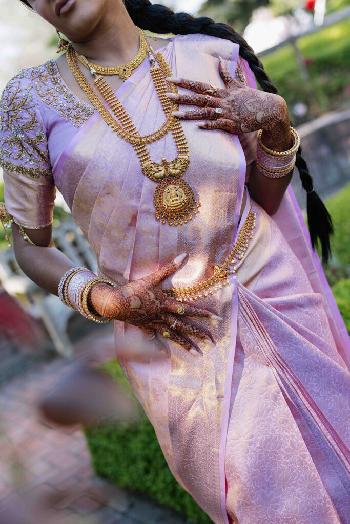 Bride poses to show off her bridal henna, layers of gold jewelry, long braid, and pink saree.