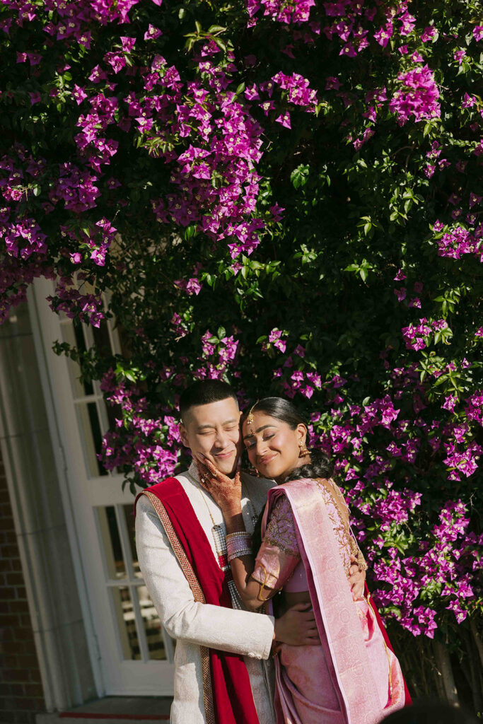 Bride grabbing grooms face as the close their eyes and smile in front of a blooming bougainvillea.