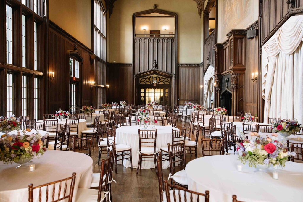 Luxury indoor wedding reception in front of a black fireplace with wood walls and round beige linen tables.