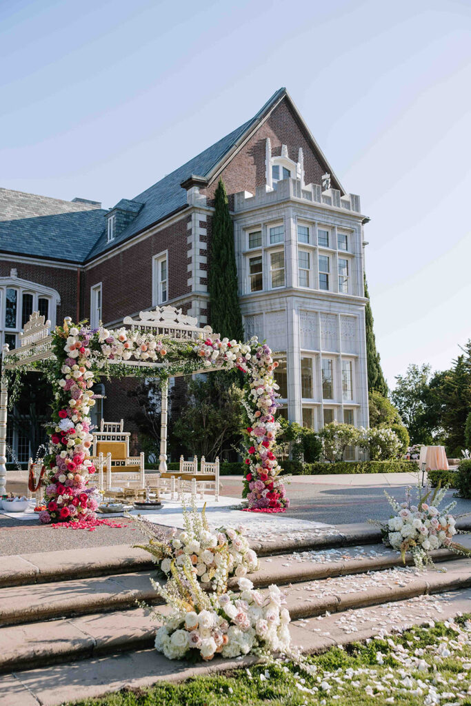 Indian mandap in white and hot pink florals with mustard yellow chairs with floral arrangements on the steps and petals leading up the aisle.