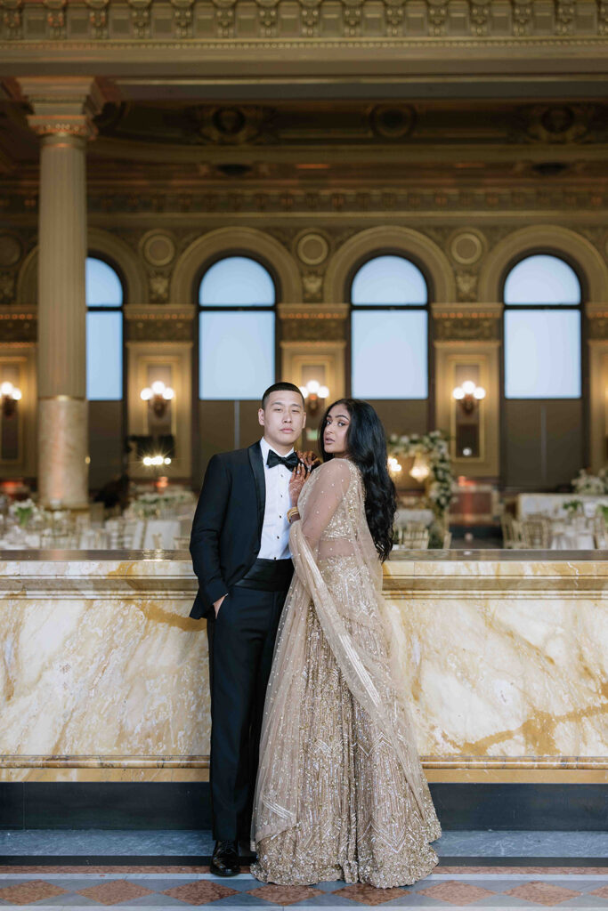 Bride in a gold bridal lehenga and groom in a tuxedo in front of their reception.