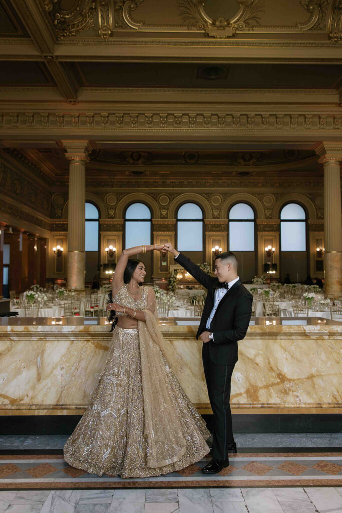 Groom, in all black suit, spinning his bride, in a gold lehenga.