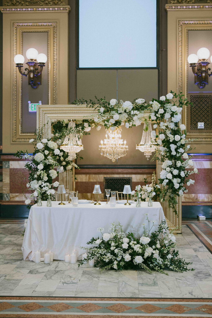 Sweetheart table with gold frame adorned with white floral arrangements and gold chandeliers hanging behind the table.