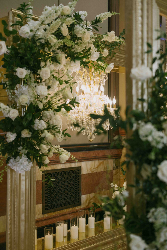 White floral arrangements hanging in front of a chandelier and candles.