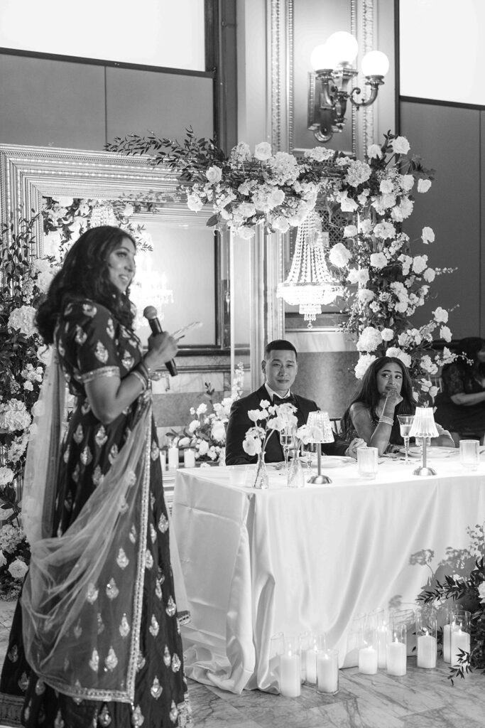 Woman giving speech in front of the sweetheart table while the couple look at her deep with emotion.