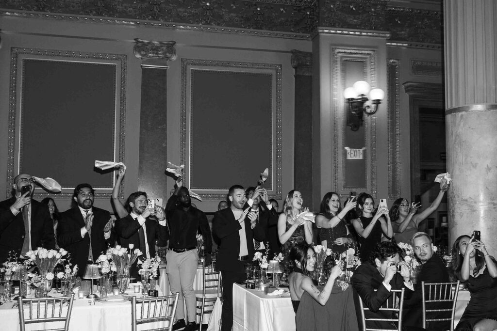 Wedding guests cheering, standing, and waving napkins in the air.