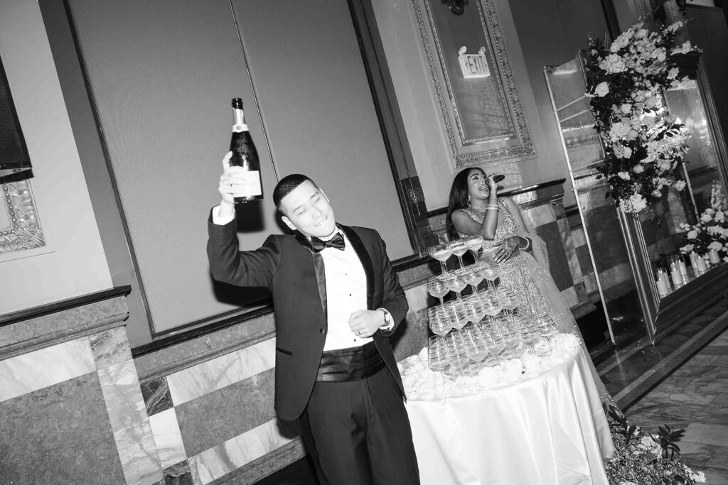 Groom holding champagne bottle up next to the champagne tower while the bride sings with a microphone.