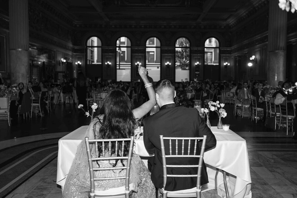 Sitting at the sweetheart table, bride raisin hand up while sitting next to groom.