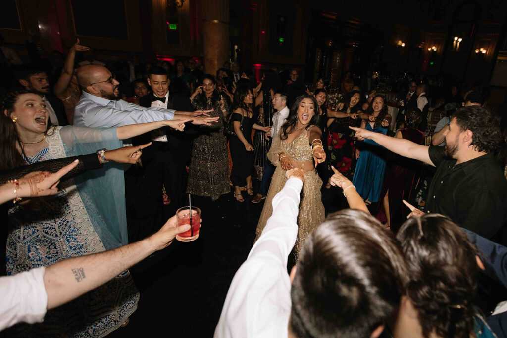 Bride in the center of the dancefloor while all her guests point at her.