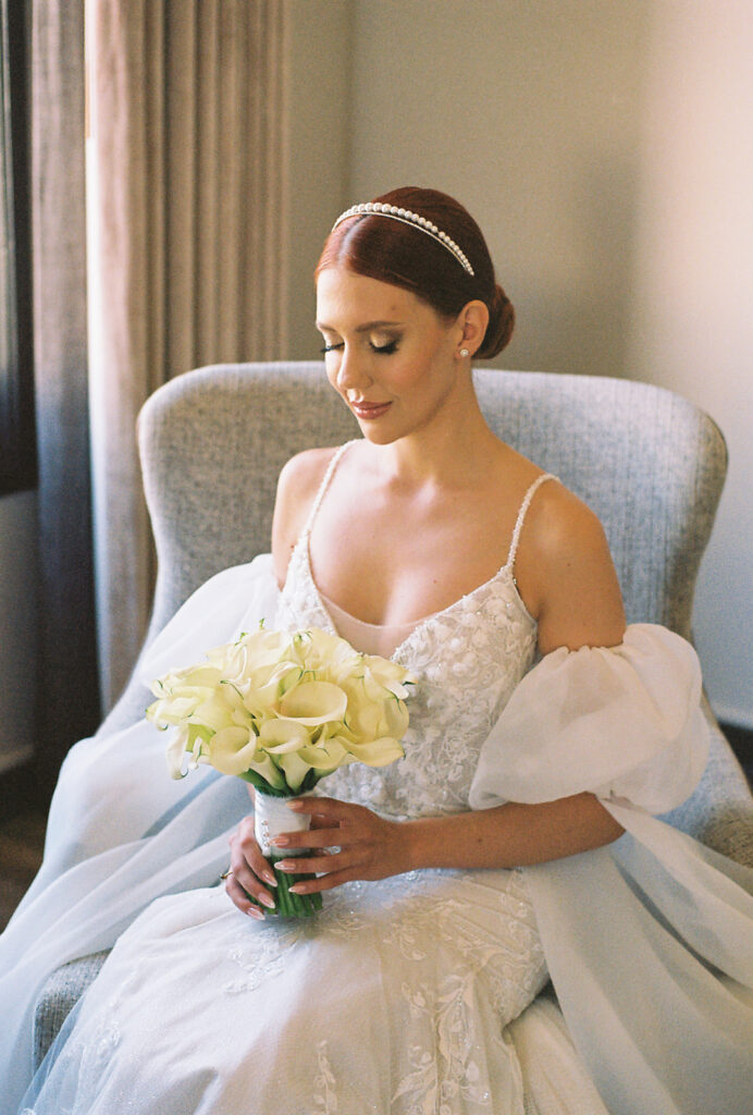 Bride looking down at her calla lily bouquet while sitting in a chair