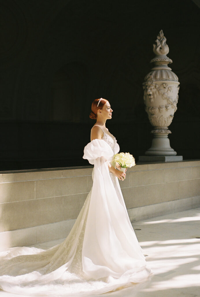 Natural light pours into city hall as bride faces windows. Her gown has a long train and she has puffy sleeves