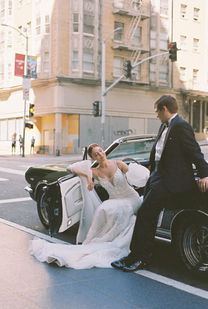 Film photo of bride sitting in driver seat with door open as groom leans on car looking down at her