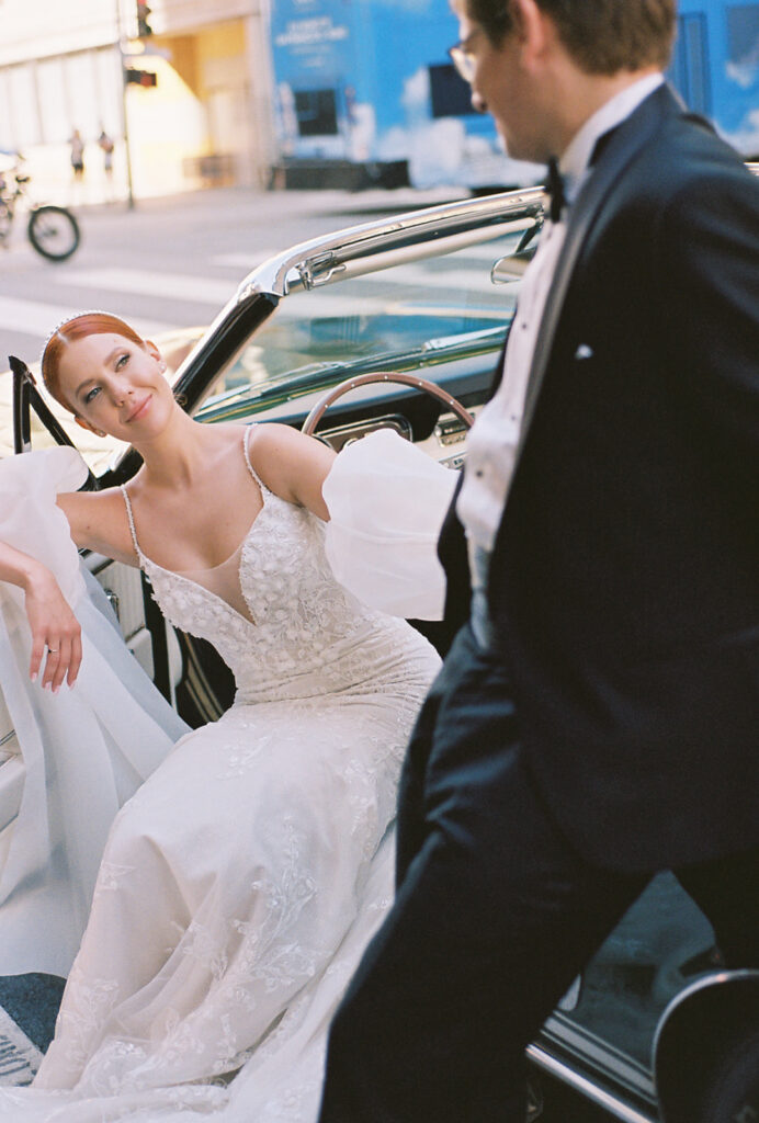 Film photo of bride sitting in driver seat with door open as groom leans on car looking down at her