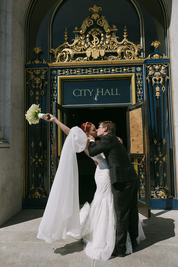 Bride holding arm and bouquet up while groom kisses her in front of entrance to city hall