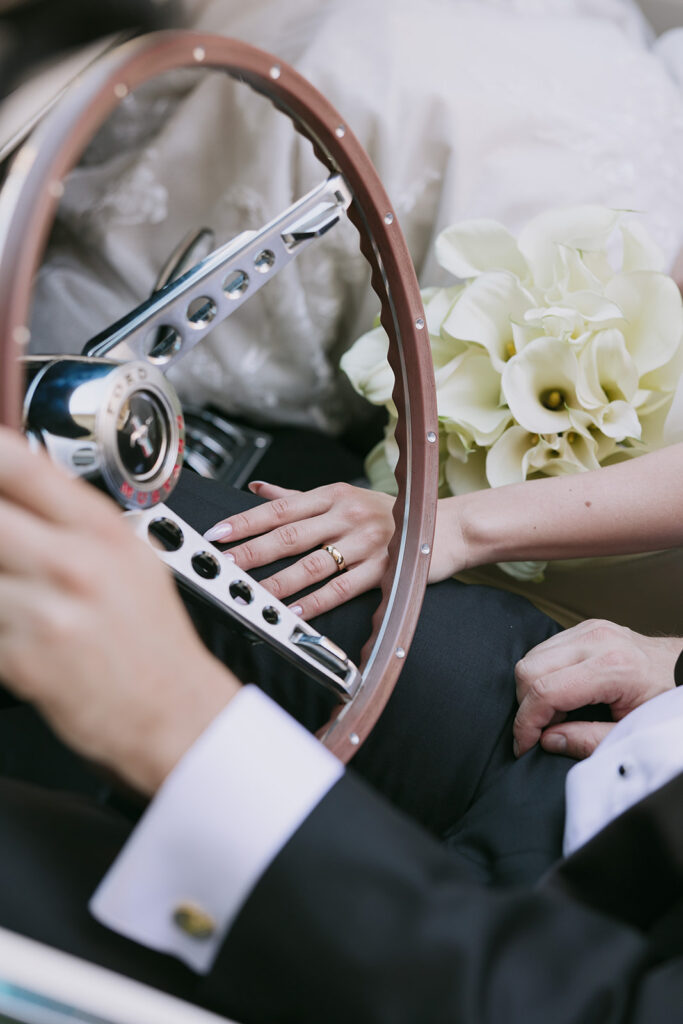 Grooms hand on steering wheel with her hand on his thigh and her bouquet in the cup holder