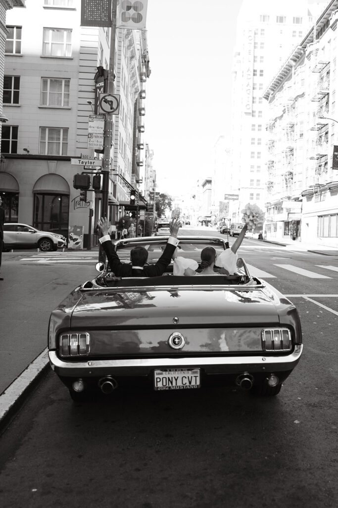Groom in drivers seat of a mustang and bride in passenger putting their hands in the air. license plate says PONYCVT