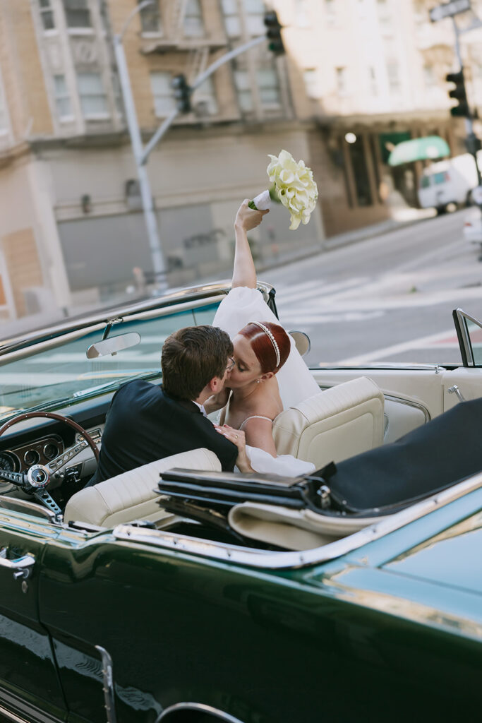 Groom in drivers seat of a mustang and bride lifting her bouquet in the air while they kiss