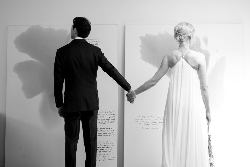 bride and groom standing far apart and holding hands in front of two art prints.