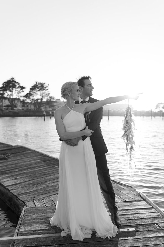Bride and groom on a dock in the bay as the sunsets behind them, bride is pointing in the direction they both are looking in. 