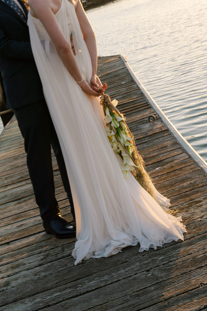 Bride and groom on a dock in the bay as the sunsets behind them, bride is holding her wrist bouquet 