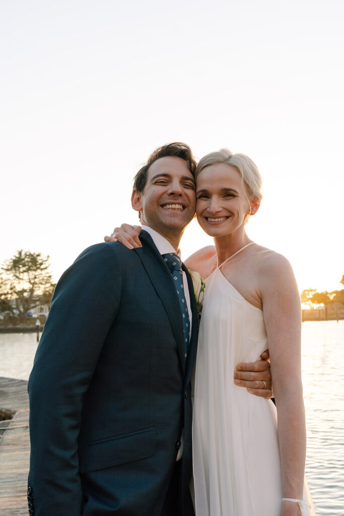 Bride and groom smiling down at the camera as the sunsets behind them