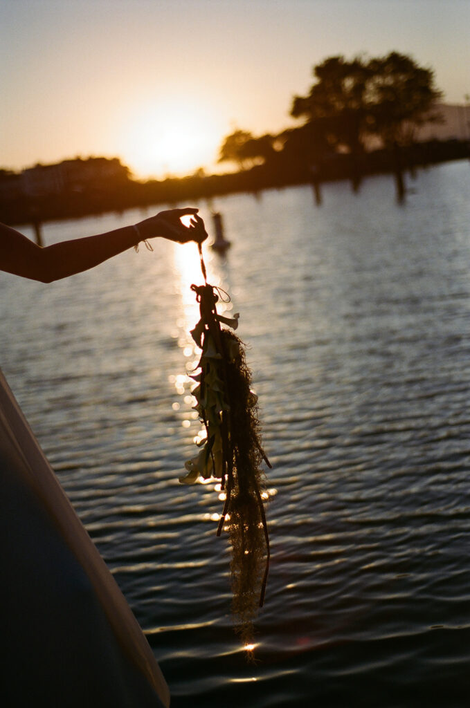 Bride is holding her wrist bouquet over the water while the sunsets behind