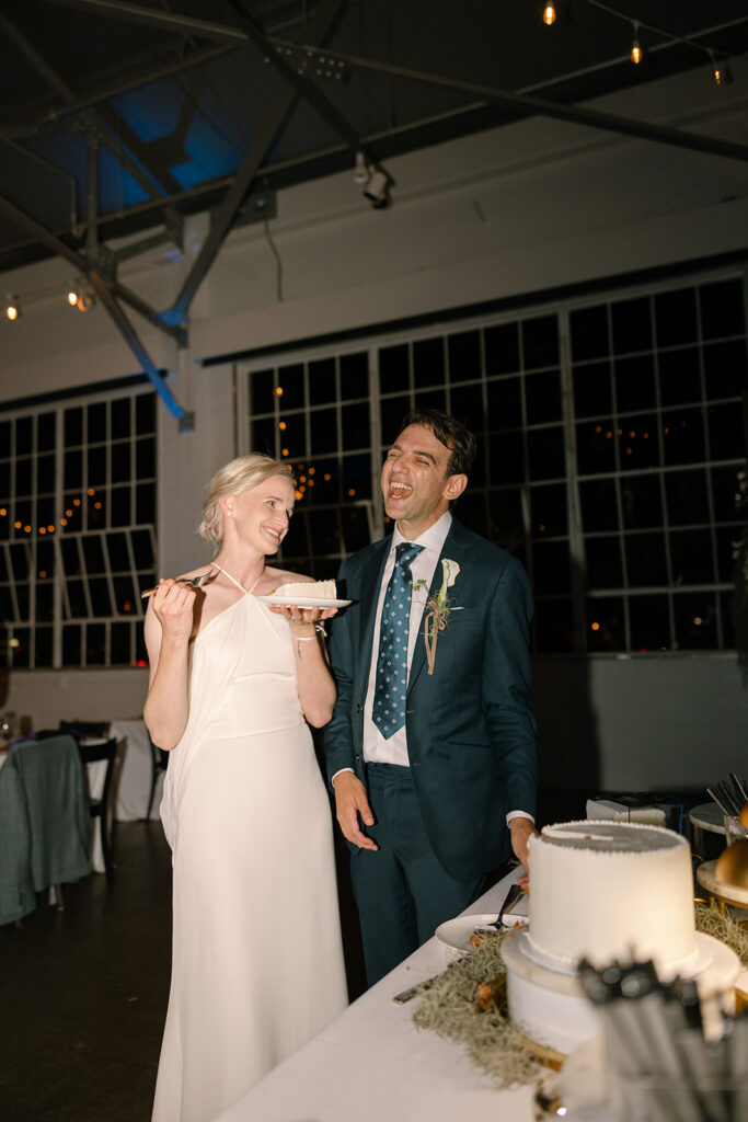 Bride and groom during their cake cutting laughing. Groom is wearing a blue tie and blue suit and bride has a bun updo