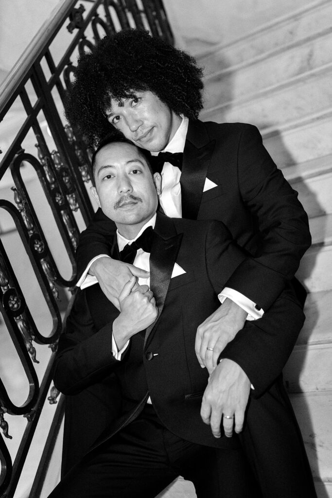 Groom leaning back into his husband on staircase embracing and looking at the camera