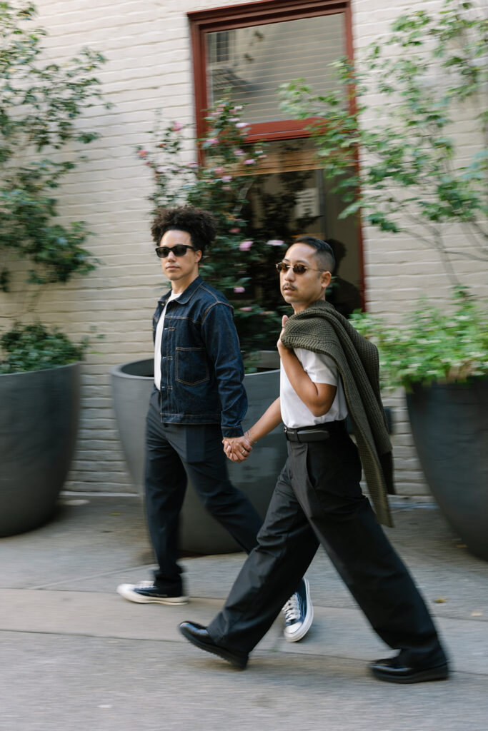 One man wearing a denim jacket, converse and trousers while the other is wearing trousers and a cardigan over his shoulder, they hold hands and are walking