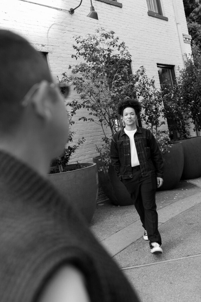 Smiling man in denim jacket and white tee walking towards and out of focus man in front of him.