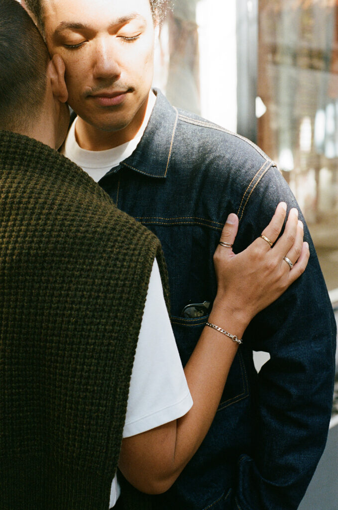 Gay couple nuzzling, man in sweater over his shoulders and layers of silver jewelry presses his hand on the man in denim jacket as his eyes are closed