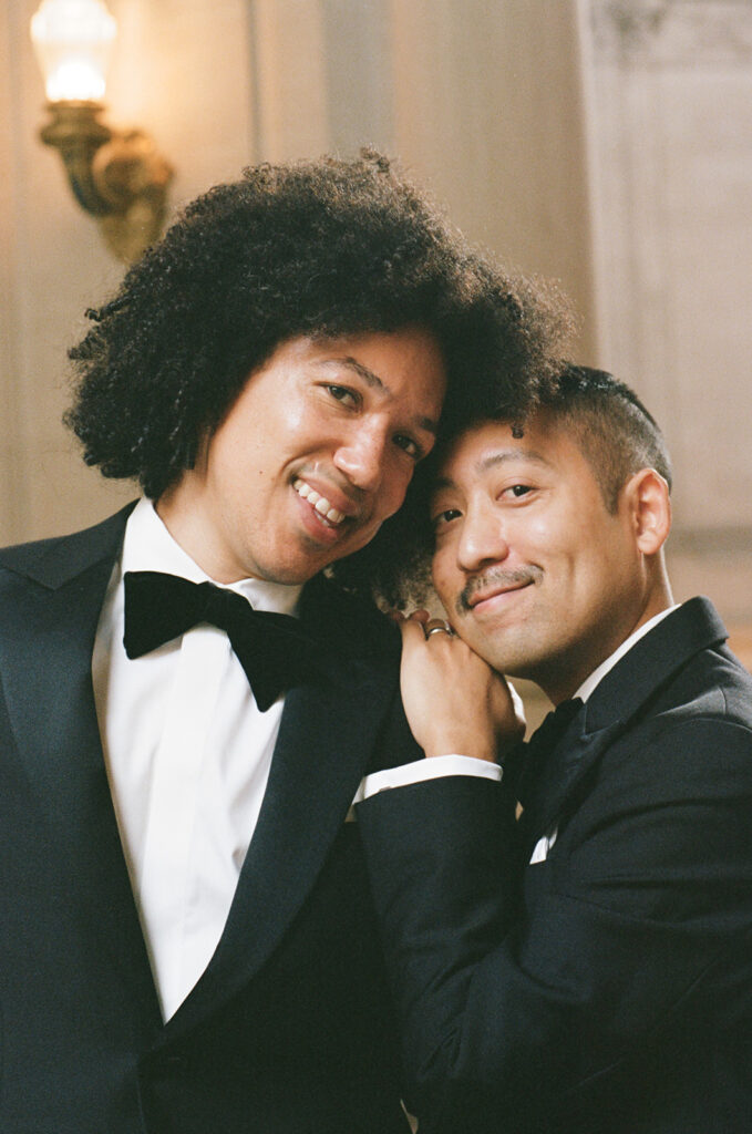 Groom leaning on his husbands shoulder while they smile at the camera