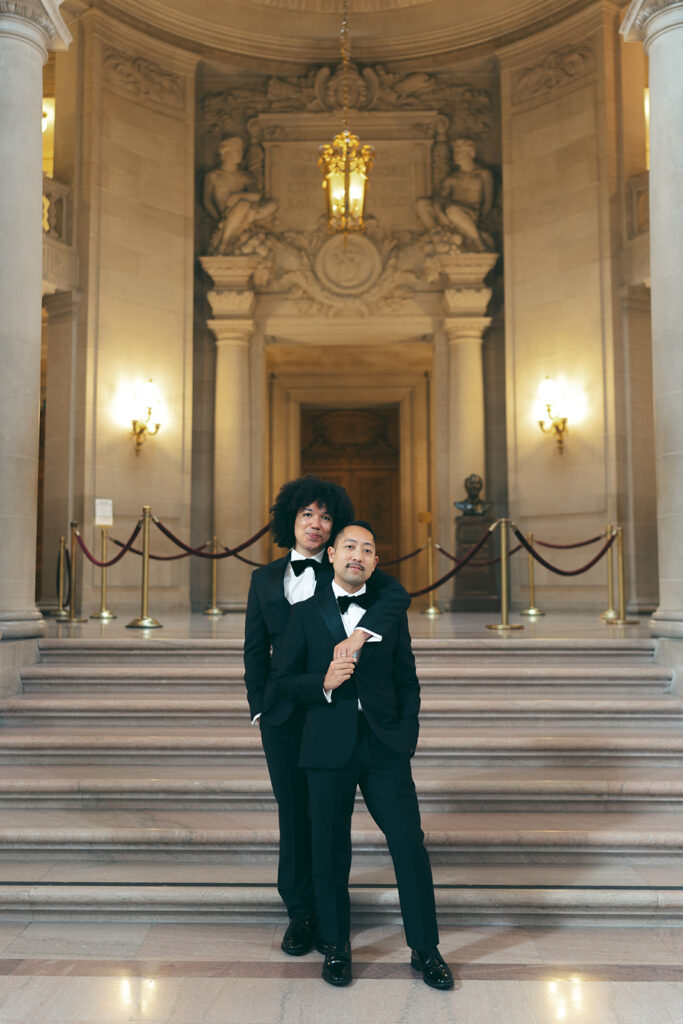 Groom slinging arm over his groom while he golds his hand standing in front of a staircase