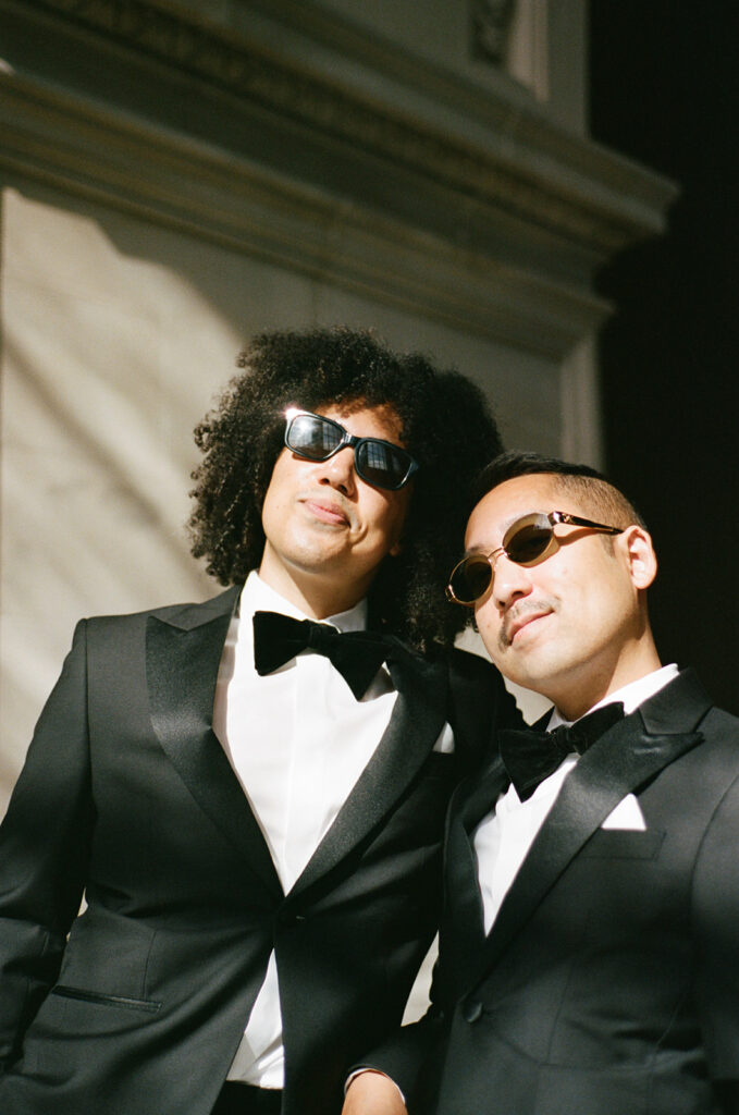 Grooms wearing sunglasses looking out the window as sun shines on them in their black tuxes with bow ties