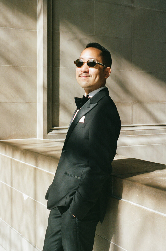 Groom wearing oval sunglasses leaning back on a ledge with his hands in his pocket