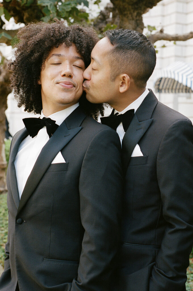 Groom kissing husbands cheek while the husband looks back and closes his eyes