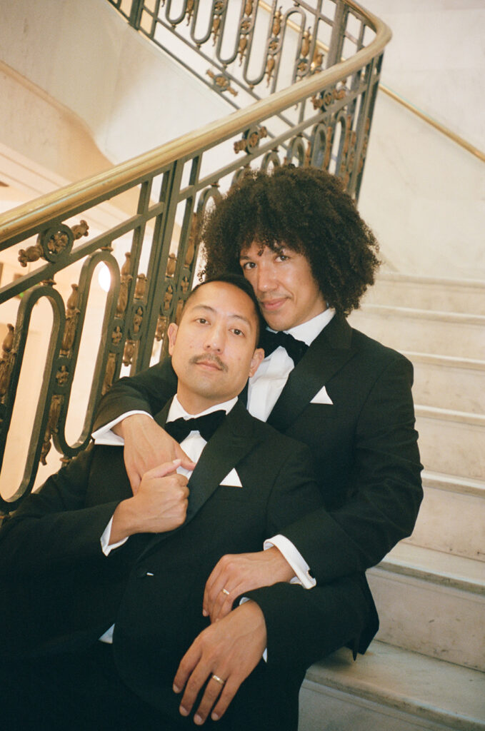 Groom leaning back into his husband on staircase embracing and looking at the camera